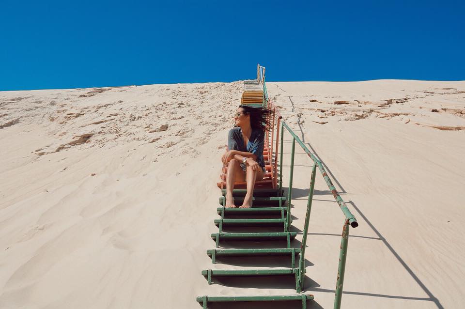 Mulher de chapéu sentada no alto de uma escada de madeira sobre uma duna de areia, sob céu azul.