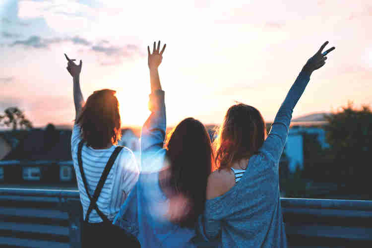 Três mulheres de costas, de braços erguidos, diante de um céu ao entardecer.
