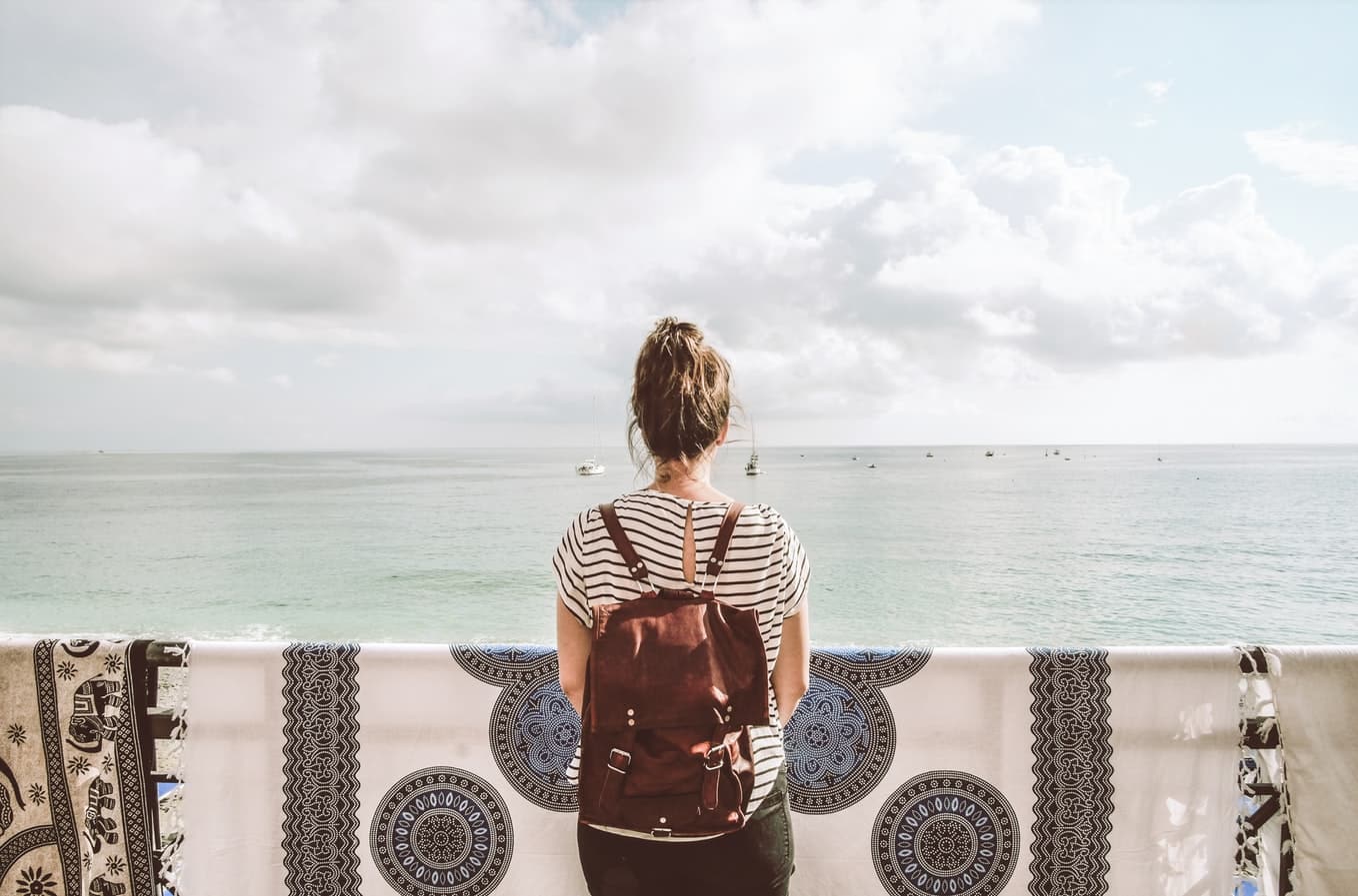Mulher de mochila, de costas, apoiada em uma sacada de azulejos, olhando para o mar.