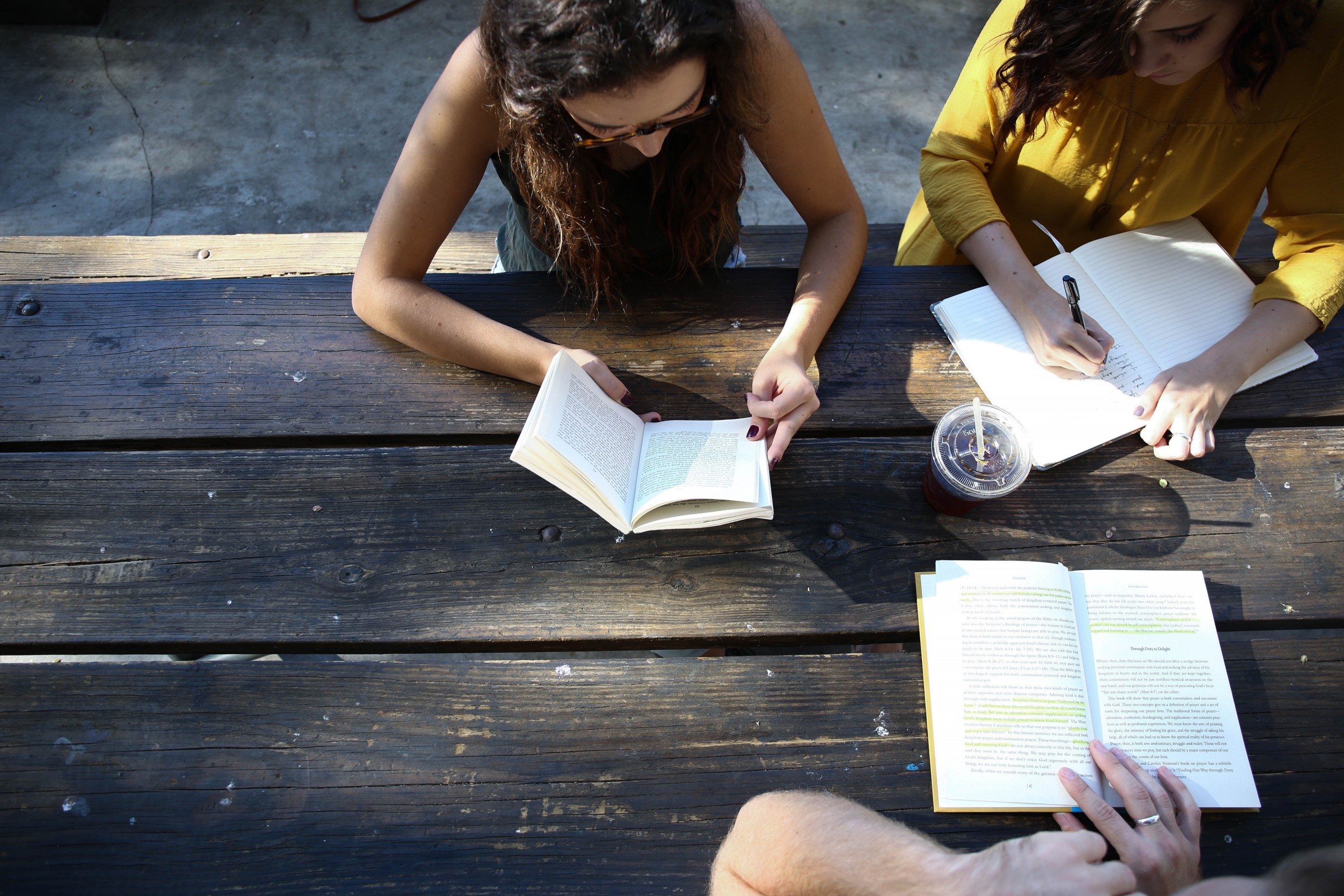 Vista de cima de três mulheres estudando com livros e cadernos abertos sobre uma mesa de madeira.