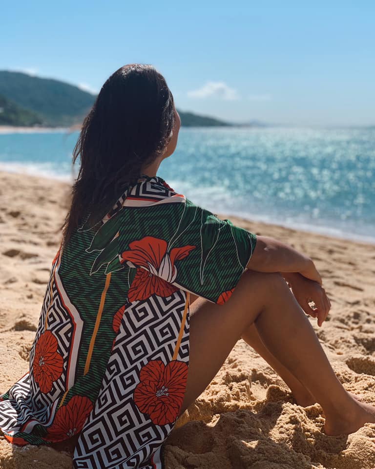 Mulher de quimono estampado sentada na areia, de costas, diante do mar.