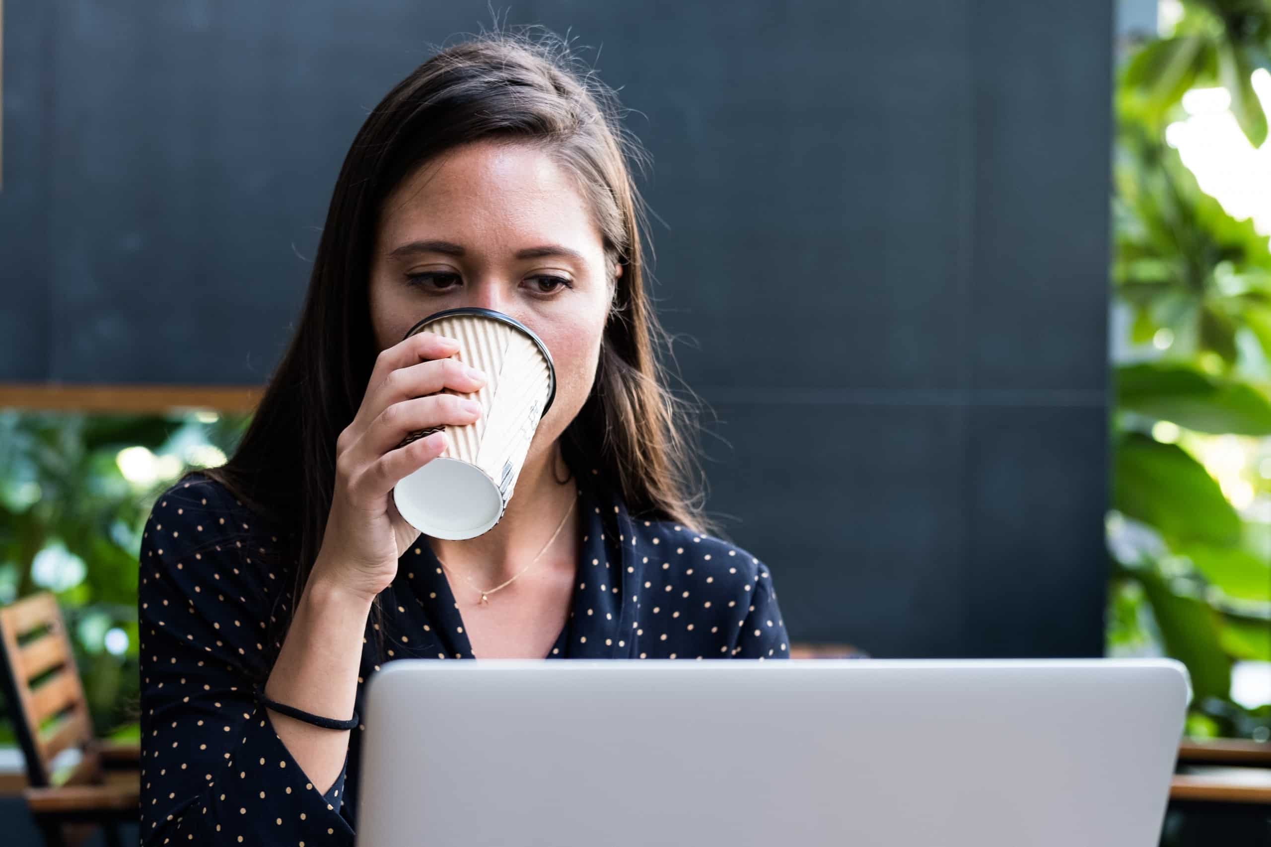Mulher bebendo café diante de um notebook, em um café cercado de plantas.