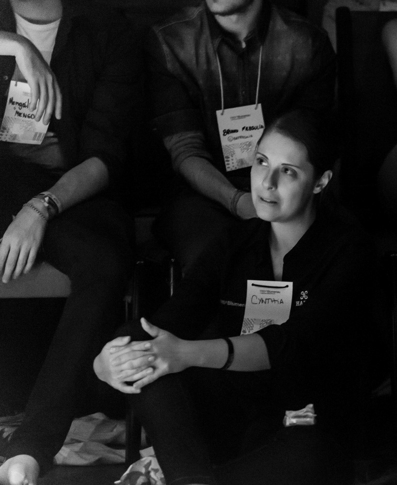 Foto em preto e branco de uma mulher de crachá sentada entre outras pessoas na plateia de um evento.