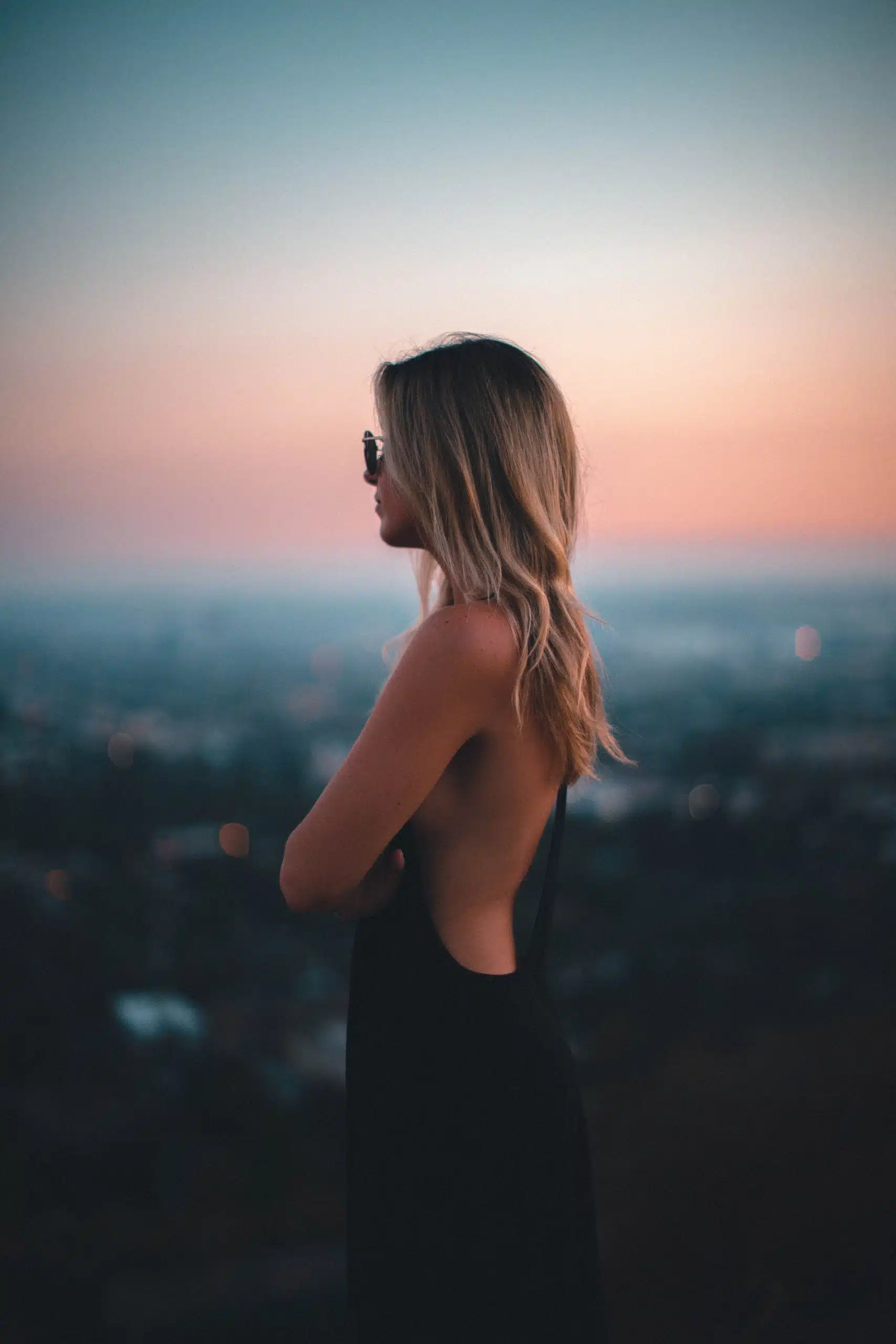 Mulher de vestido escuro, de costas, diante de uma paisagem urbana ao entardecer.