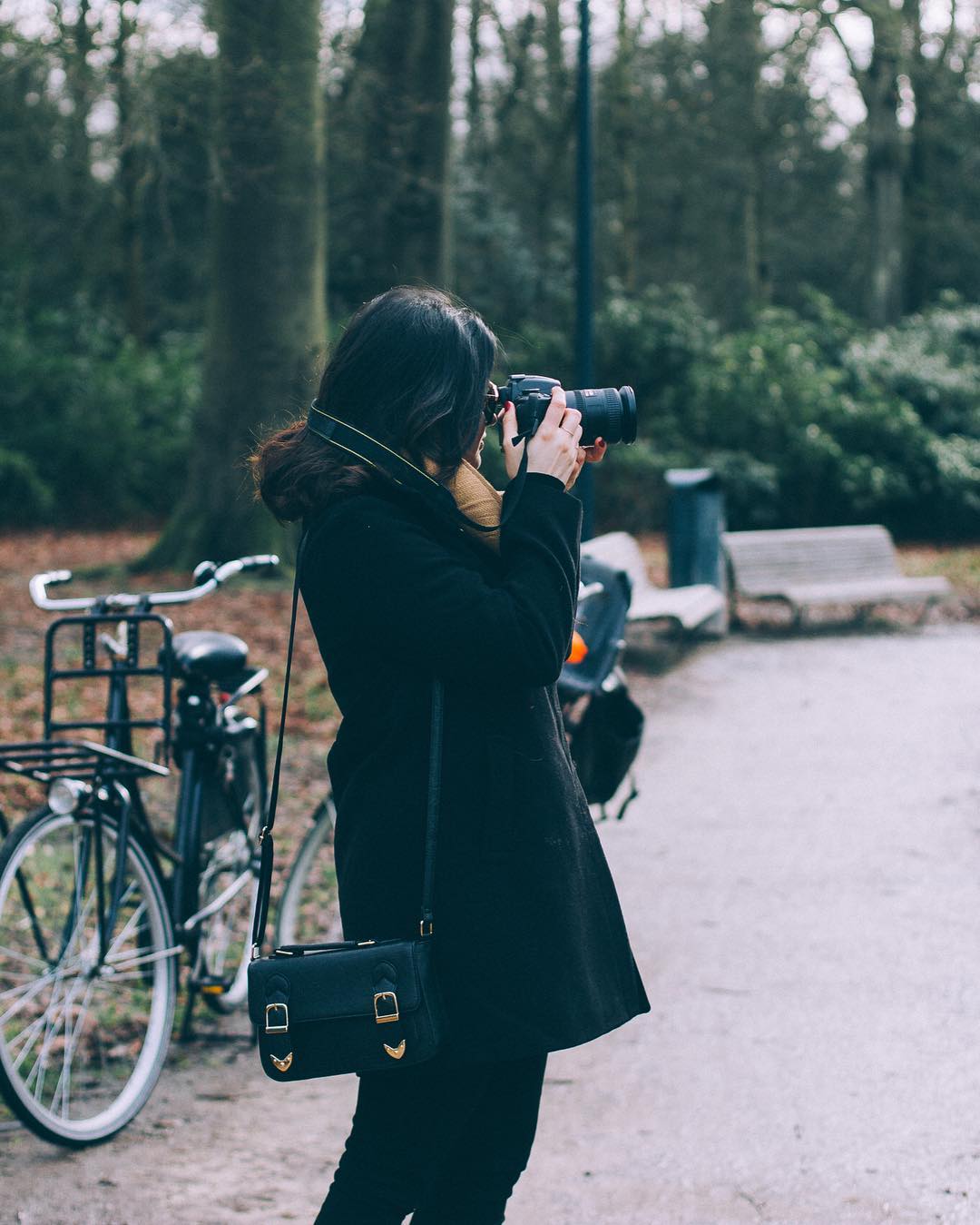 Mulher de casaco preto fotografando com uma câmera ao lado de uma bicicleta, em um parque arborizado.