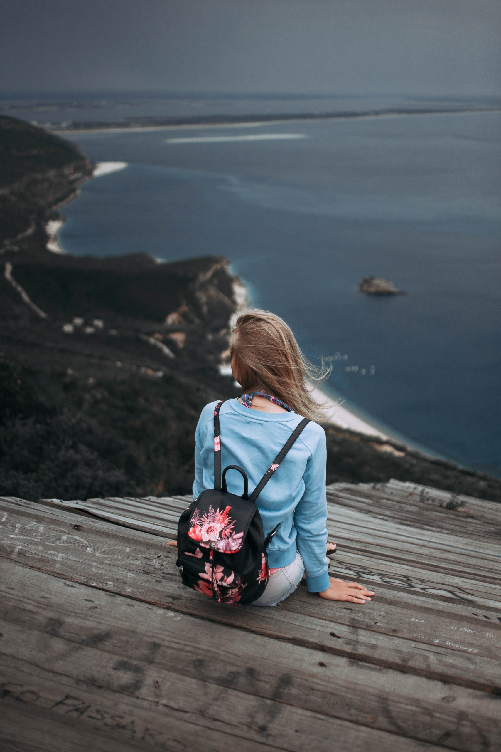 Mulher com mochila sentada em um deque de madeira, de costas, diante de uma enseada ao longe.